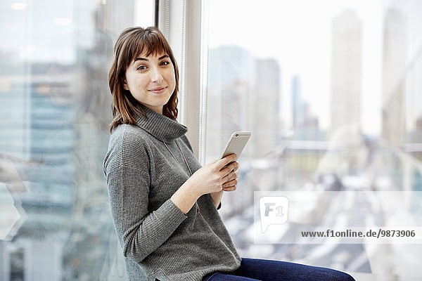 A woman holding a smart phone seated at a window with a city view.