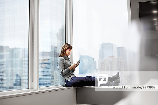 Eine Frau  die ein Smartphone in der Hand hält und auf einem Fensterbrett sitzt.