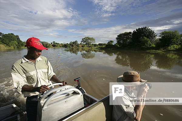 'Man with camera and tour guide driving boat down a tranquil river in Lower Zambezi National Park; Zambia'