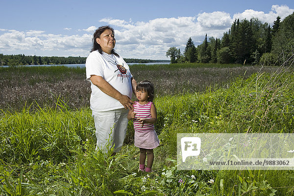 Native Aboriginal Grandmother Standing In A Field Beside A Lake With Her Granddaughter In Shoal Lake  Ontario  Canada.