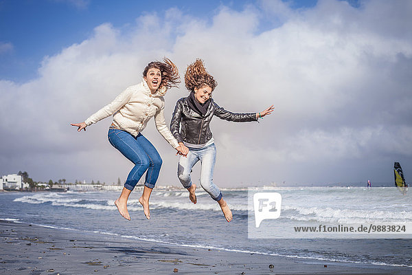 Zwei junge Frauen  die Händchen halten und am Strand springen.