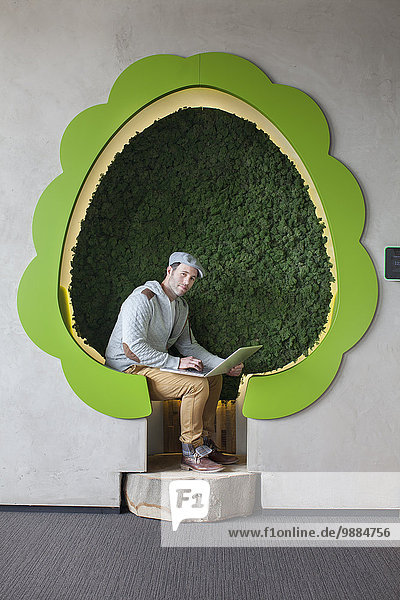 Portrait eines männlichen Designers bei der Arbeit am Laptop in baumförmigen Büroräumen