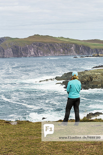 Woman standing on grassy cliff overlooking rugged shoreline with waves; Ballinglanna  County Cork  Ireland