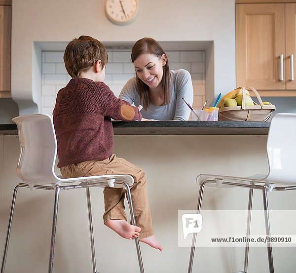 Boy sitting on stool with mother in kitchen and drawing