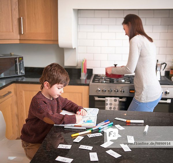Junge färbt im Buch auf der Küchenzeile  während die Mutter das Abendessen zubereitet.