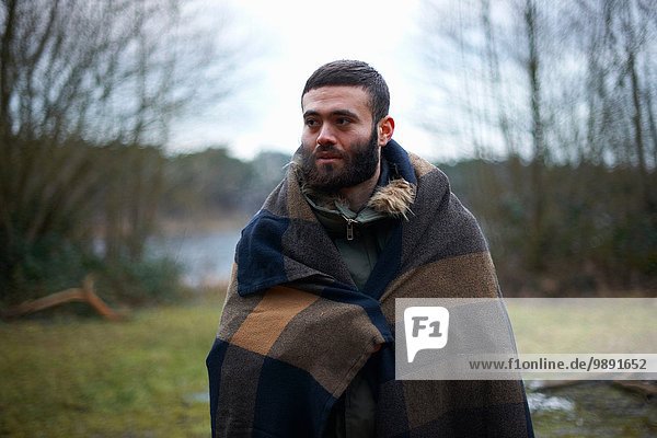 Young male camper wrapped in blanket in woods