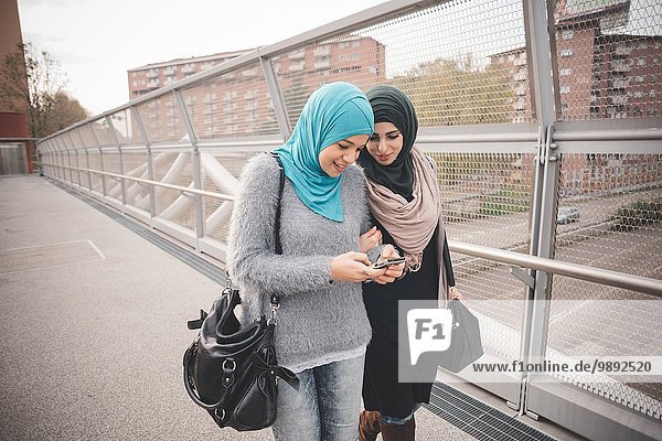 Two female friends on footbridge reading smartphone texts