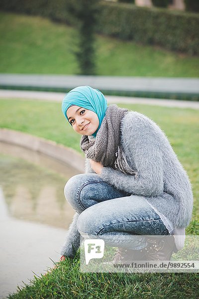 Porträt einer jungen Frau mit Hijab  die sich am Parksee hockt