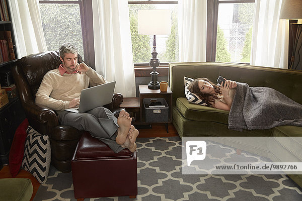 Young couple in sitting room using smartphone and laptop