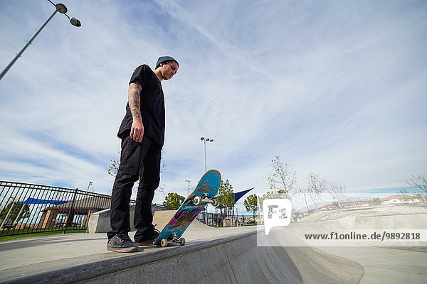 Young male skateboarder standing on edge of skateboard park