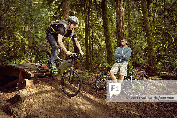 Young man riding mountain bike over ramp  in forest  while friend watches