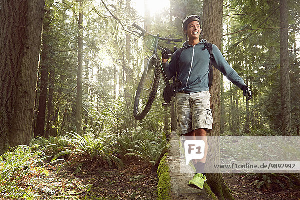 Junger Mann im Wald  trägt Mountainbike über umgestürztem Baum