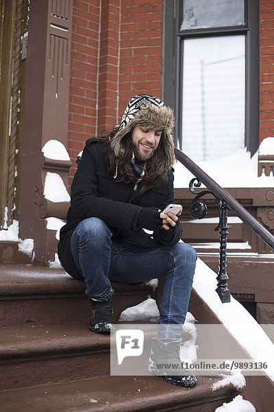 Junger Mann liest Text auf Smartphone auf schneebedeckter Haustürstufe