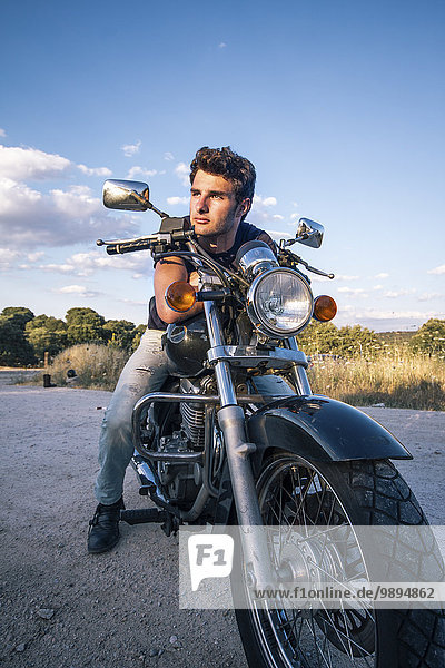 Spanien  Madrid  junger Mann  der sich auf seinem Motorrad auf einer unbefestigten Straße bei Sunse lehnt
