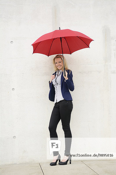 Lächelnde Geschäftsfrau mit rotem Regenschirm an Betonwand gelehnt