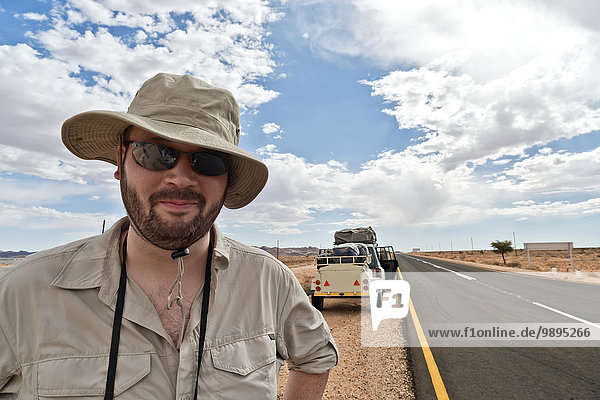 Namibia  Mann mit Safarihut und Sonnenbrille mit Auto am Straßenrand im Hintergrund