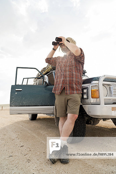 Namibia  Etosha Nationalpark  Mann  der sich auf das Auto stützt und durch ein Fernglas schaut.