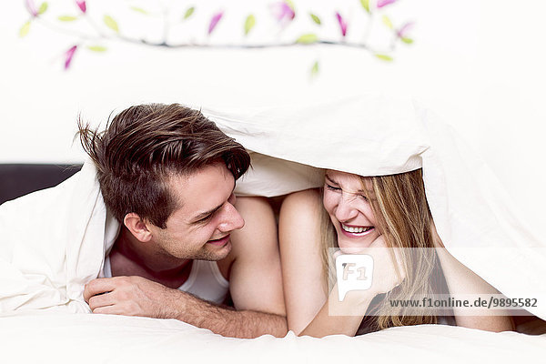 Laughing couple lying side by side in bed under blanket
