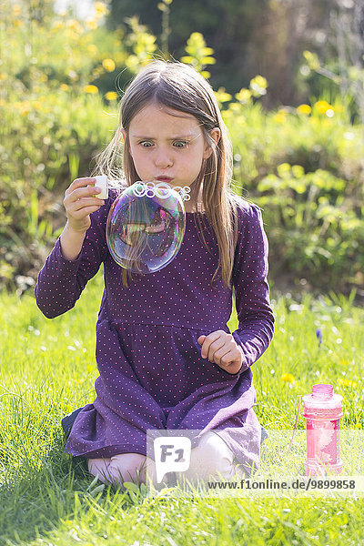 Mädchen hockt auf einer Wiese und bläst große Seifenblase.