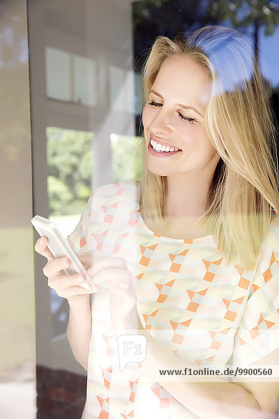 Blonde Frau mit Smartphone am Fenster stehend