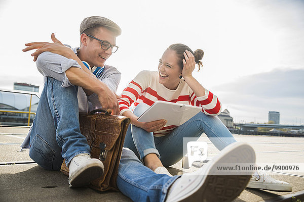 Happy young man and woman sitting on ground using digital tablet