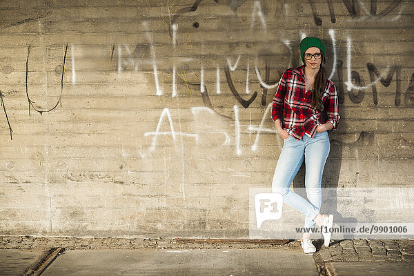 Junge Frau mit kariertem Hemd und Wollmütze an Graffiti-Wand lehnend