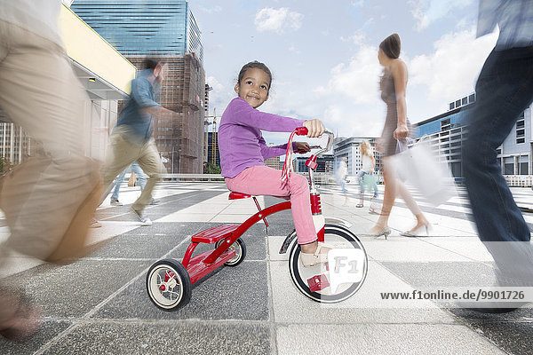 Girl riding a tricycle between a crowd of people in a city