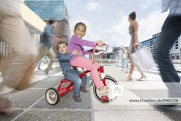 Mädchen und Junge auf einem Dreirad zwischen einer Menge von Menschen in einer Stadt.