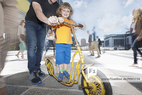 Vater hilft seinem Sohn  einen Roller zu fahren zwischen einer Menschenmenge in einer Stadt.