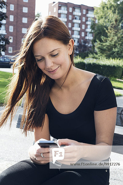Lächelnde junge Frau mit Smartphone auf der Bank im Freien