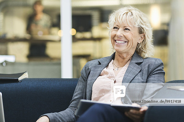 Fröhliche Geschäftsfrau schaut weg  während sie das digitale Tablett auf dem Sofa in der Bürolobby hält.