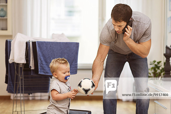 Vater benutzt das Handy  während er dem Baby zu Hause den Ball gibt.