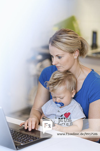 Mutter arbeitet am Laptop  während das Baby das Handy am Tisch benutzt.