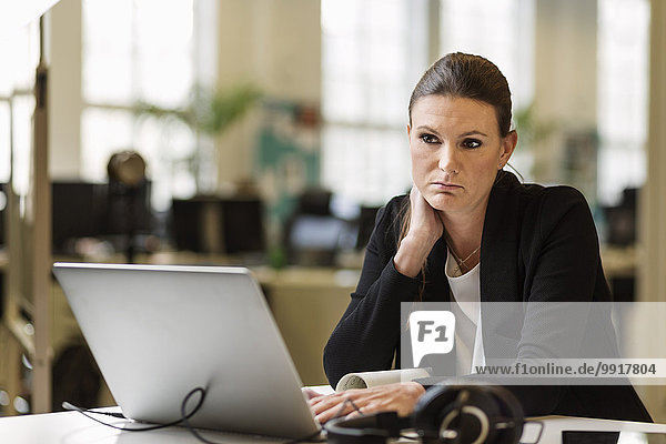 Nachdenkliche Geschäftsfrau schaut weg  während sie den Laptop am Schreibtisch im Kreativbüro benutzt.
