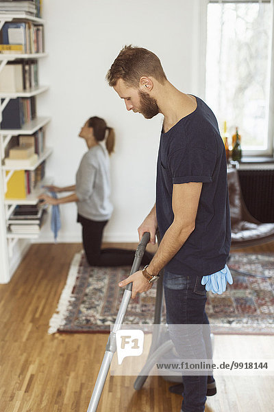Seitenansicht des Mannes beim Staubsaugen des Bodens,  während die Frau zu Hause im Hintergrund Regale putzt.