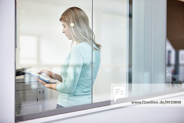 Mittlere erwachsene Geschäftsfrau mit digitalem Tablett hinter dem Bürofenster