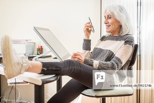 Porträt einer älteren Frau  die am Computer sitzt und ein Smartphone mit Fuß auf dem Schreibtisch hält.