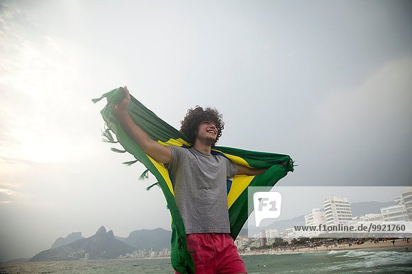 Lächelnder junger Mann mit brasilianischer Flagge am Strand von Ipanema  Rio De Janeiro  Brasilien