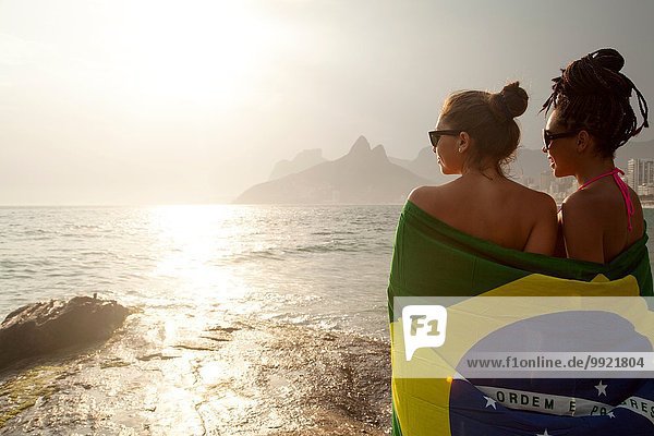 Rear view of two young women wrapped in Brazilian flag  Ipanema beach  Rio De Janeiro  Brazil
