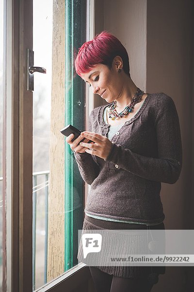 Junge Frau steht am Fenster  mit dem Smartphone