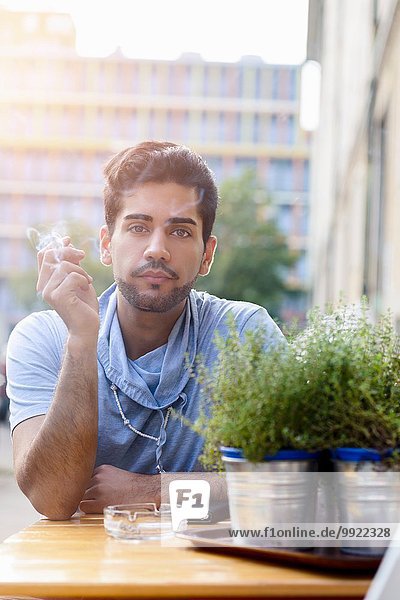 Porträt eines jungen Mannes im Freien  am Tisch sitzend  Zigarette rauchend
