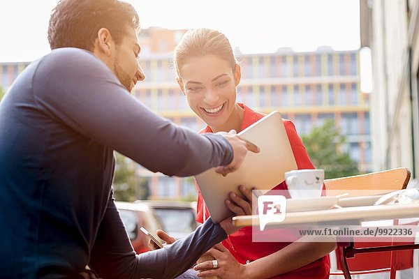 Junger Mann und junge Frau sitzen vor dem Café und schauen auf das digitale Tablett.