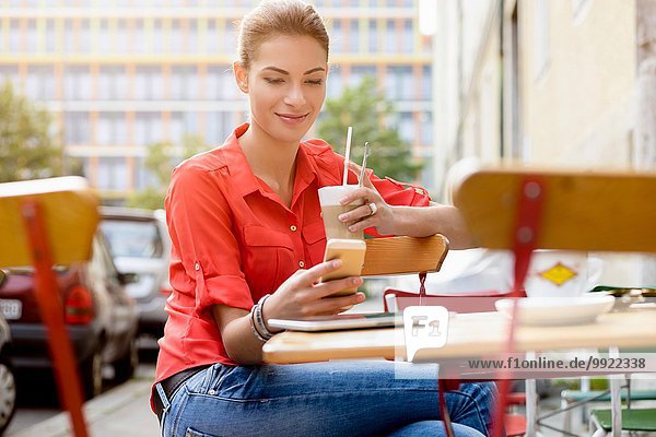 Junge Frau sitzend vor dem Café mit Getränk  mit Handy