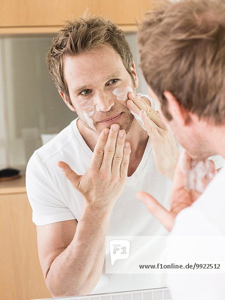 Mann  der in den Badezimmerspiegel schaut und Gesichtscreme aufträgt.
