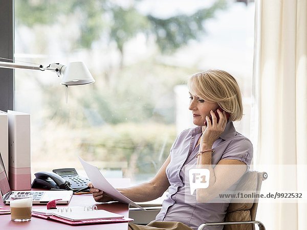 Reife Geschäftsfrau im Gespräch auf dem Smartphone am heimischen Schreibtisch