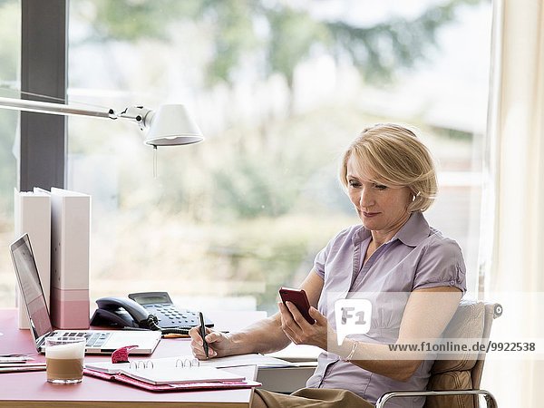 Mature businesswoman reading text message on smartphone at home desk