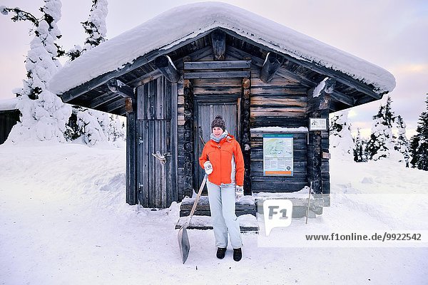 Porträt einer jungen Frau mit Schneeschaufel vor dem Blockhaus  Posio  Lappland  Finnland