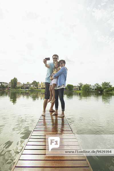 Family with young daughter standing together at end of lake pier taking selfie
