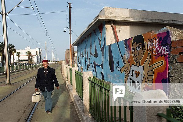 Man wearing a traditional tarbouche walking along a wall covered with graffiti in Tunis.