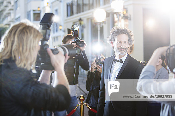 Smiling celebrity in tuxedo being photographed by paparazzi at red carpet event